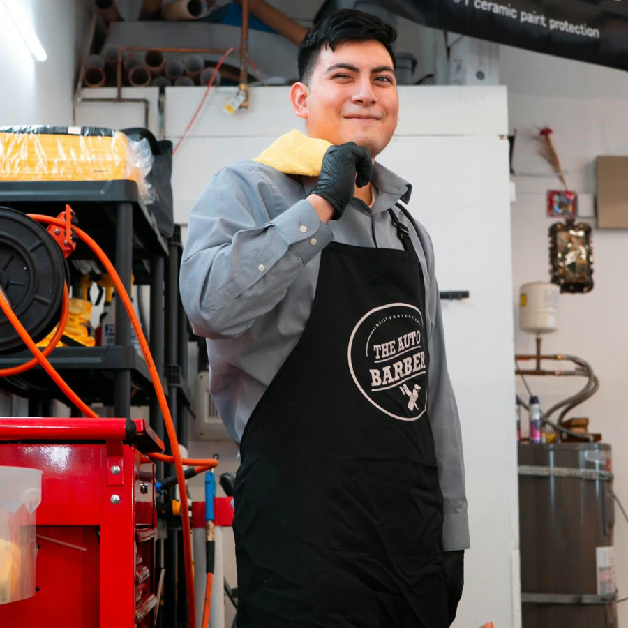 Hikari in the Auto Barber shop, wearing the branded apron and holding a detailing towel with a gloved hand